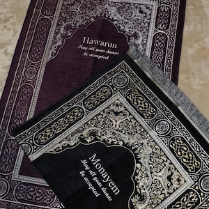 Op de afbeelding: Twee gebedskleden, &eacute;&eacute;n donkerpaars en &eacute;&eacute;n zwart, met sierlijke zilveren en gouden ontwerpen. Het paarse kleed heeft de tekst "Hawarun, May all your duas be accepted." Het zwarte kleed heeft de tekst "Monayem, May all your duas be accepted."