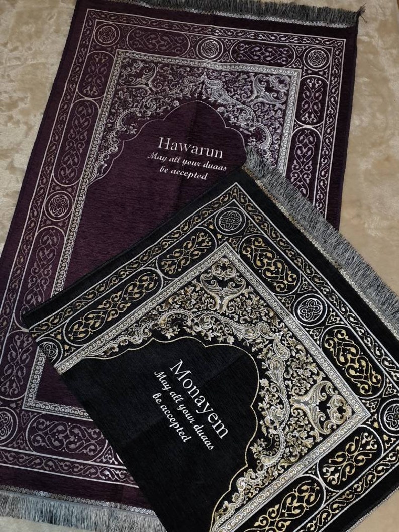 Op de afbeelding: Twee gebedskleden, &eacute;&eacute;n donkerpaars en &eacute;&eacute;n zwart, met sierlijke zilveren en gouden ontwerpen. Het paarse kleed heeft de tekst "Hawarun, May all your duas be accepted." Het zwarte kleed heeft de tekst "Monayem, May all your duas be accepted."