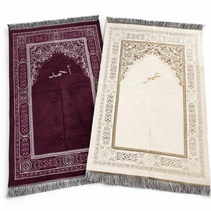 Op de afbeelding: Twee gebedskleden met sierlijke ontwerpen en gefranjerde randen. E&eacute;n is diep bordeauxrood met witte accenten en Arabisch schrift. De andere is gebroken wit met gouden accenten en Arabisch schrift. Beide hebben een centraal boogontwerp.