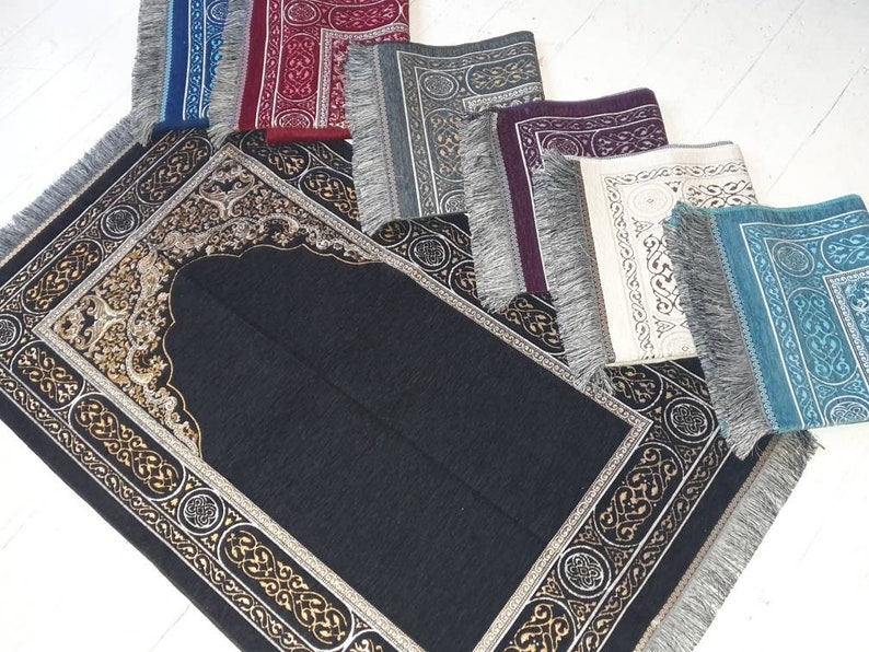 Op de afbeelding: Een verzameling gebedskleden in diverse kleuren, waaronder zwart, blauw, rood, grijs, paars, wit en turquoise. Elk kleed heeft een sierlijk randontwerp met gouden accenten en een franjerand. De kleden zijn op een wit oppervlak geplaatst.