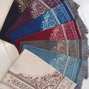 Op de afbeelding: Een verzameling gebedskleden in diverse kleuren, waaronder zwart, bordeauxrood, blauw en cr&egrave;me. Elk kleed heeft een sierlijke, patroonrijke rand en een centraal ontwerp. De kleden hebben gefranjerde randen en zijn in een waaier gerangschikt.