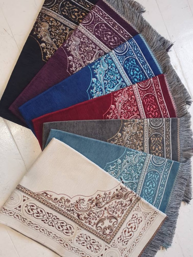 Op de afbeelding: Een verzameling gebedskleden in diverse kleuren, waaronder zwart, bordeauxrood, blauw en cr&egrave;me. Elk kleed heeft een sierlijke, patroonrijke rand en een centraal ontwerp. De kleden hebben gefranjerde randen en zijn in een waaier gerangschikt.