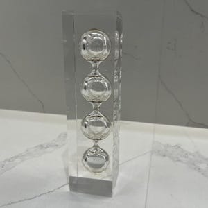 May include: A clear, rectangular glass object containing four silver-colored spheres stacked vertically. The spheres are connected by narrow glass stems. The object is displayed on a white surface with a gray background.