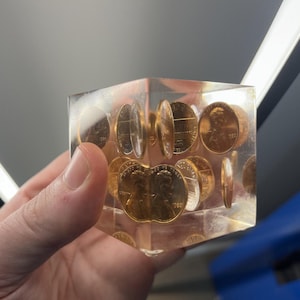 May include: A clear resin cube encasing multiple copper pennies. The pennies are arranged throughout the cube, showcasing the heads and tails of the coins. The cube is held in a hand.