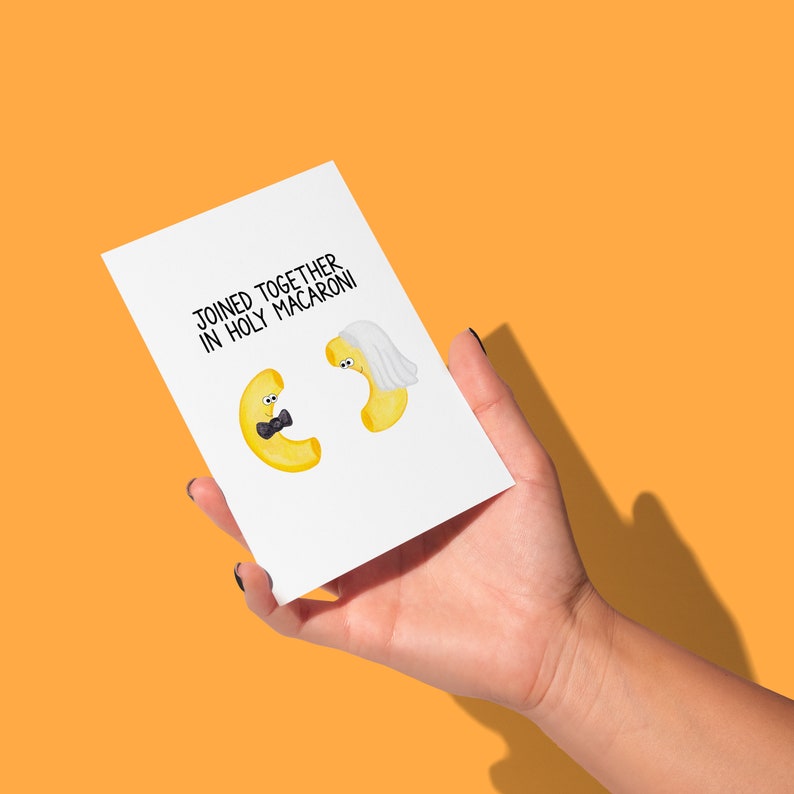 Op de afbeelding: Een witte wenskaart met de tekst "JOINED TOGETHER IN HOLY MACARONI" toont cartoon macaroni karakters. E&eacute;n macaroni heeft een vlinderdas, de andere draagt een sluier. De kaart wordt tegen een oranje achtergrond gehouden.