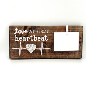 以下が含まれることがあります： 「Love at First Heartbeat」の白い文字が入った濃い茶色の木製サイン。 文字の下には、ストリングアートのハートと心拍のデザインがあります。 白いクリップが白いカードを保持しています。