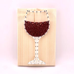 May include: A red wine glass made with string art on a wooden board. The glass is filled with red string and the stem and base are white string.