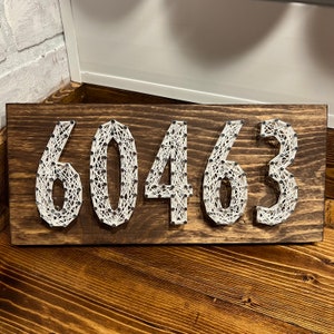 May include: A wooden sign with the numbers 60463 crafted from white string and small black nails. The numbers are displayed on a dark brown, rectangular wooden board. The sign is a decorative piece.
