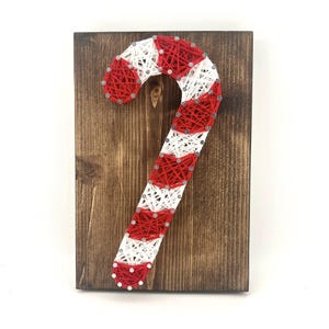 May include: A handcrafted Christmas decoration featuring a candy cane design made with red and white string art on a stained wooden board. The candy cane is secured with small silver nails, creating a festive holiday accent.