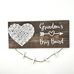 May include: A wooden "Grandma's Brag Board" with a white string art heart and text. The dark brown board has a jute string with four small white clothespins for displaying photos or notes. A charming gift.