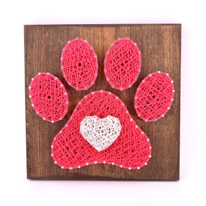 May include: A wooden wall art piece featuring a paw print design made with pink string. A white heart is in the center of the paw. The artwork is mounted on a dark brown wooden board, creating a striking contrast.