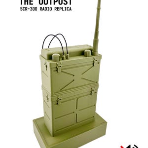 May include: An olive-green SCR-300 radio replica, labeled "The Outpost." The radio has a telescoping antenna, two black wire loops, and a rectangular base. The text "DISPLAY ONLY - NO AUDIO" is also visible.