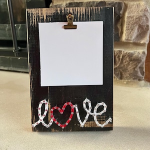May include: A rustic wooden picture frame with a black finish. The frame holds a white card secured by a metal clip. The word "love" is spelled out in red and white string art across the bottom of the frame.