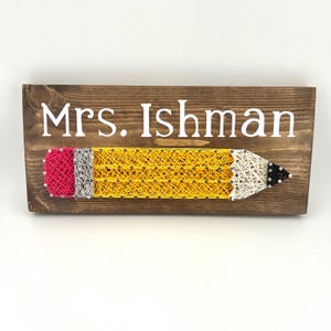 Puede incluir: Letrero de madera con el nombre "Mrs. Ishman" en letras blancas. Un lápiz de arte de hilo amarillo, rojo, plateado y negro aparece debajo del nombre. El letrero está hecho de madera marrón oscuro.