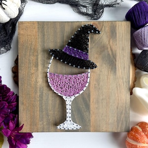 May include: A string art piece on a square wooden board featuring a wine glass filled with light purple string and topped with a black witch hat that has a purple band. The design is created using silver nails and colored string.