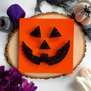 May include: A square orange wooden board featuring a jack-o'-lantern face created with black nails and black string. The design includes two triangular eyes, a triangular nose, and a wide, jagged mouth formed by the intricate string art pattern.