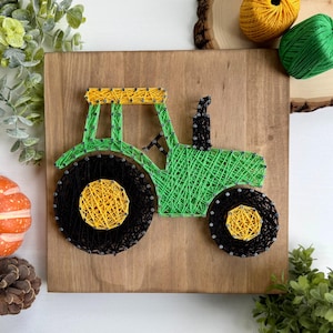 Op de afbeelding: Een draadkunstwerk op een vierkant houten bord dat een tractor voorstelt. De tractor is gemaakt met groene, zwarte en gele draad gewikkeld rond metalen spijkers. Het ontwerp bevat een groot zwart achterwiel, een kleiner voorwiel en een groene carrosserie.