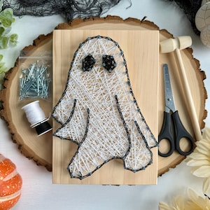 May include: A DIY string art kit featuring a rectangular light wood board with a ghost design outlined in black nails and filled with white string. The kit includes a bag of silver nails, a spool of white string, a small wooden hammer, and scissors.