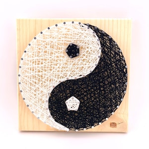 May include: A wooden board with a black and white string art design of the yin yang symbol. The symbol is made with nails and string, and the background is a natural wood color.