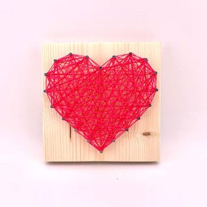 May include: A pink heart-shaped string art piece on a wooden background. The heart is made of string wrapped around nails hammered into the wood.