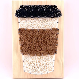 May include: A wooden board with a string art design of a coffee cup. The cup is made of brown, white, and black string wrapped around nails hammered into the wood.