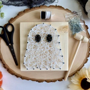 May include: A DIY string art kit featuring a ghost design created with white string on a square wooden board, two black string eyes, a small wooden hammer, a bag of silver nails, and two spools of black and white thread for assembly.