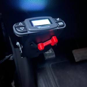 May include: A black electronic device with a digital display and buttons. The device has a red toggle switch. The device is mounted in a vehicle.