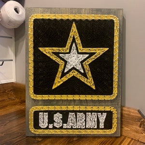 May include: Wooden string art featuring the U.S. Army logo. The design includes a large gold star over a black background, with a white star in the center. The words "U.S. ARMY" are displayed in white string below the star.