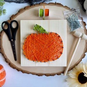 May include: A square wooden board featuring a string art pumpkin design created with orange and green thread wrapped around silver nails. The kit includes black scissors, a small wooden hammer, a plastic bag of silver nails, and two thread spools.