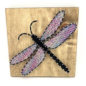 May include: A handcrafted string art dragonfly on a wooden board. The dragonfly features a black body and wings filled with colorful threads in shades of pink, purple, blue, and white. The wings are outlined with small silver nails.