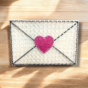 May include: A handmade string art piece in the shape of an envelope with a pink heart. The envelope is made of white string, with a black border and diagonal lines. The heart is centered and made of pink string.