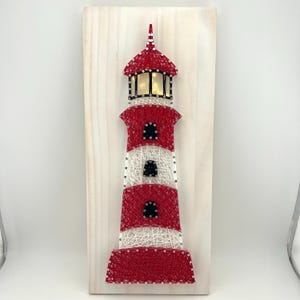 May include: A string art lighthouse with red and white detailing. The lighthouse is mounted on a light-colored wooden board. The lighthouse features a red roof and red and white stripes. The lighthouse has a light inside.