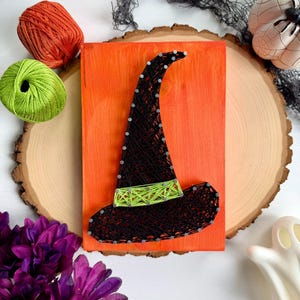 May include: A rectangular orange wooden board featuring a string art design of a witch hat. The hat is created with black string and silver nails, with a bright lime green string band around the base. The design is centered on the board.