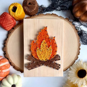 May include: A rectangular wooden plaque featuring a string art design of a campfire. The flames are created with orange and yellow thread wrapped around silver nails, while the logs at the base are formed with brown thread on the light wood surface.