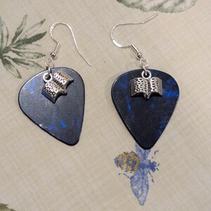 Puede incluir: Pendientes con púas de guitarra negras con motas azules. Cada púa tiene un pequeño dije plateado con forma de libro abierto. Los pendientes tienen ganchos para las orejas plateados.