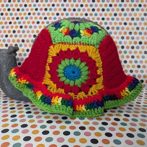 May include: A colorful crocheted snail shell with red, yellow, green, and blue yarn. The shell is a crocheted granny square pattern with a flower design in the center.