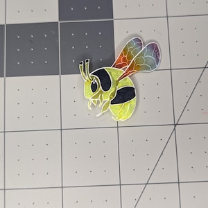 Może przedstawiać: Kolorowa naklejka z pszczołą z żółtymi, czarnymi i tęczowymi skrzydłami. Pszczoła jest przedstawiona w stylu kreskówkowym, ze szczegółowym wzorem. Naklejka jest umieszczona na szarej i białej powierzchni kratki.