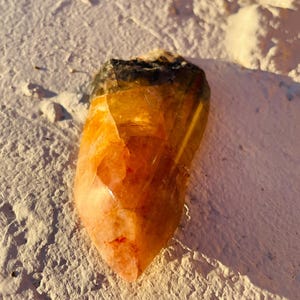 May include: A raw, natural citrine crystal with a gradient of colors, from a dark, almost black base to a translucent, golden-orange tip. The crystal is resting on a textured, off-white surface.