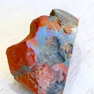 May include: A rough-hewn, natural stone with a mix of colors. The stone features a blend of red, gray, and blue hues, with a polished, reflective surface on one side. The overall shape is irregular, showcasing the stone's natural form.