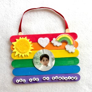 May include: A rainbow-colored popsicle stick craft with a photo of a child, a felt sun, a felt heart, and a felt rainbow. The bottom row of popsicle sticks has the words "You Are My Sunshine" spelled out with letter beads.