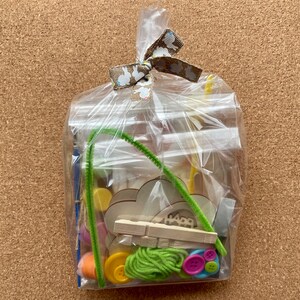以下が含まれることがあります： ウサギ柄のリボンで結ばれた透明なセロハンバッグ。クラフト用品が入っています。木製の形、ボタン、モール、糸が含まれています。「Happy」の文字が木製の形に表示されています。