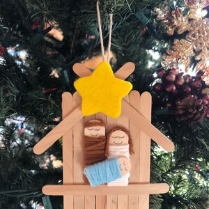 May include: A Christmas bauble made from craft sticks, yarn, and felt. It depicts the nativity scene with Mary, Joseph, and baby Jesus in a manger. A yellow felt star is on top of the manger.