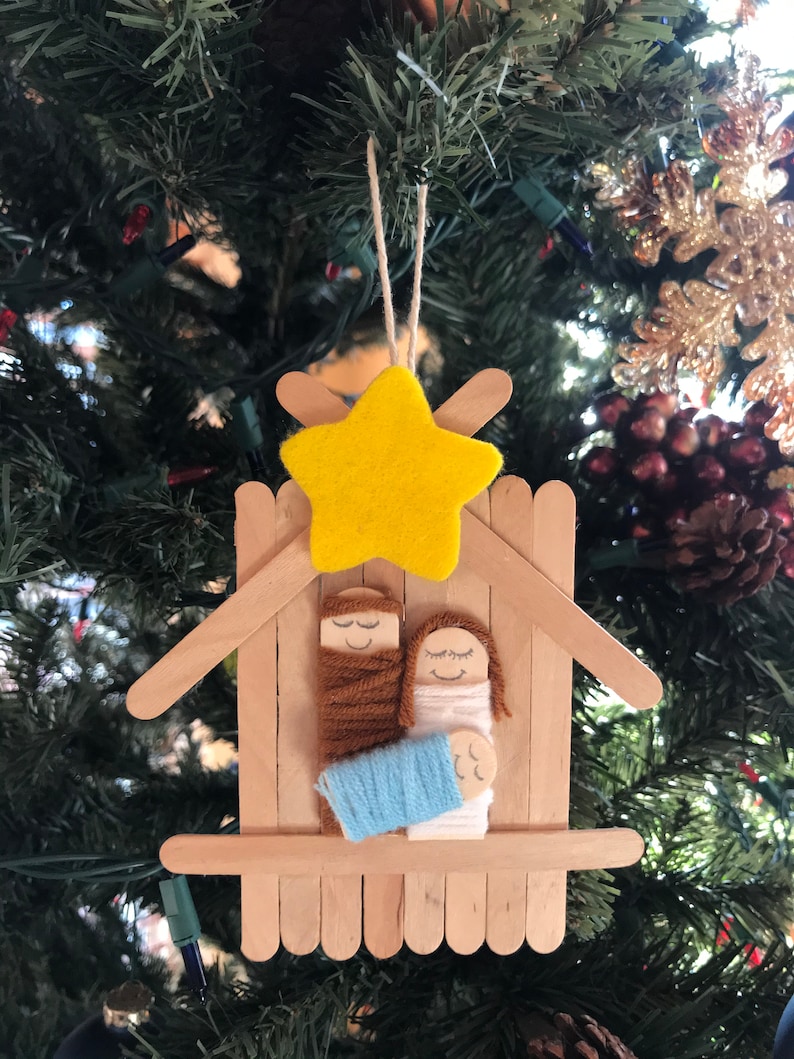 May include: A Christmas bauble made from craft sticks, yarn, and felt. It depicts the nativity scene with Mary, Joseph, and baby Jesus in a manger. A yellow felt star is on top of the manger.
