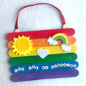 May include: A rainbow-colored popsicle stick sign with a felt sun, rainbow, and heart. The sign says "You are my sunshine" in white letters on blue circles.