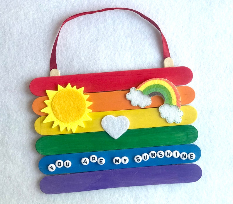 May include: A rainbow-colored popsicle stick sign with a felt sun, rainbow, and heart. The sign says "You are my sunshine" in white letters on blue circles.
