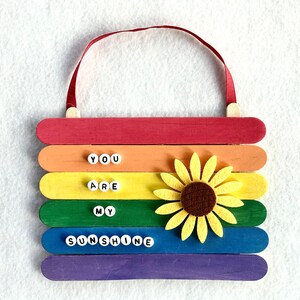 May include: A rainbow-colored wooden sign with the words "You are my sunshine" spelled out in white letters. A yellow felt sunflower is attached to the sign.