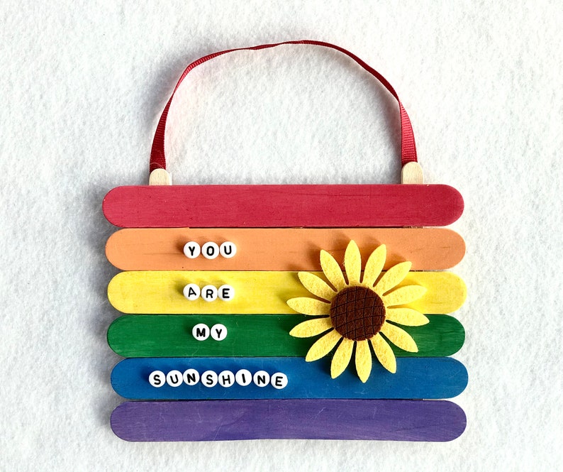 May include: A rainbow-colored wooden sign with the words "You are my sunshine" spelled out in white letters. A yellow felt sunflower is attached to the sign.