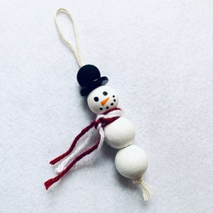 Può includere: Ciondolo a forma di pupazzo di neve bianco con un cappello a cilindro nero, una sciarpa rossa e bianca e un viso dipinto.