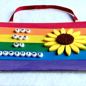 May include: A rainbow-colored wooden sign with the words "You are my sunshine" spelled out in white beads. A felt sunflower is attached to the sign.