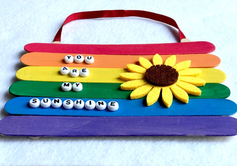 May include: A rainbow-colored wooden sign with the words "You are my sunshine" spelled out in white beads. A felt sunflower is attached to the sign.